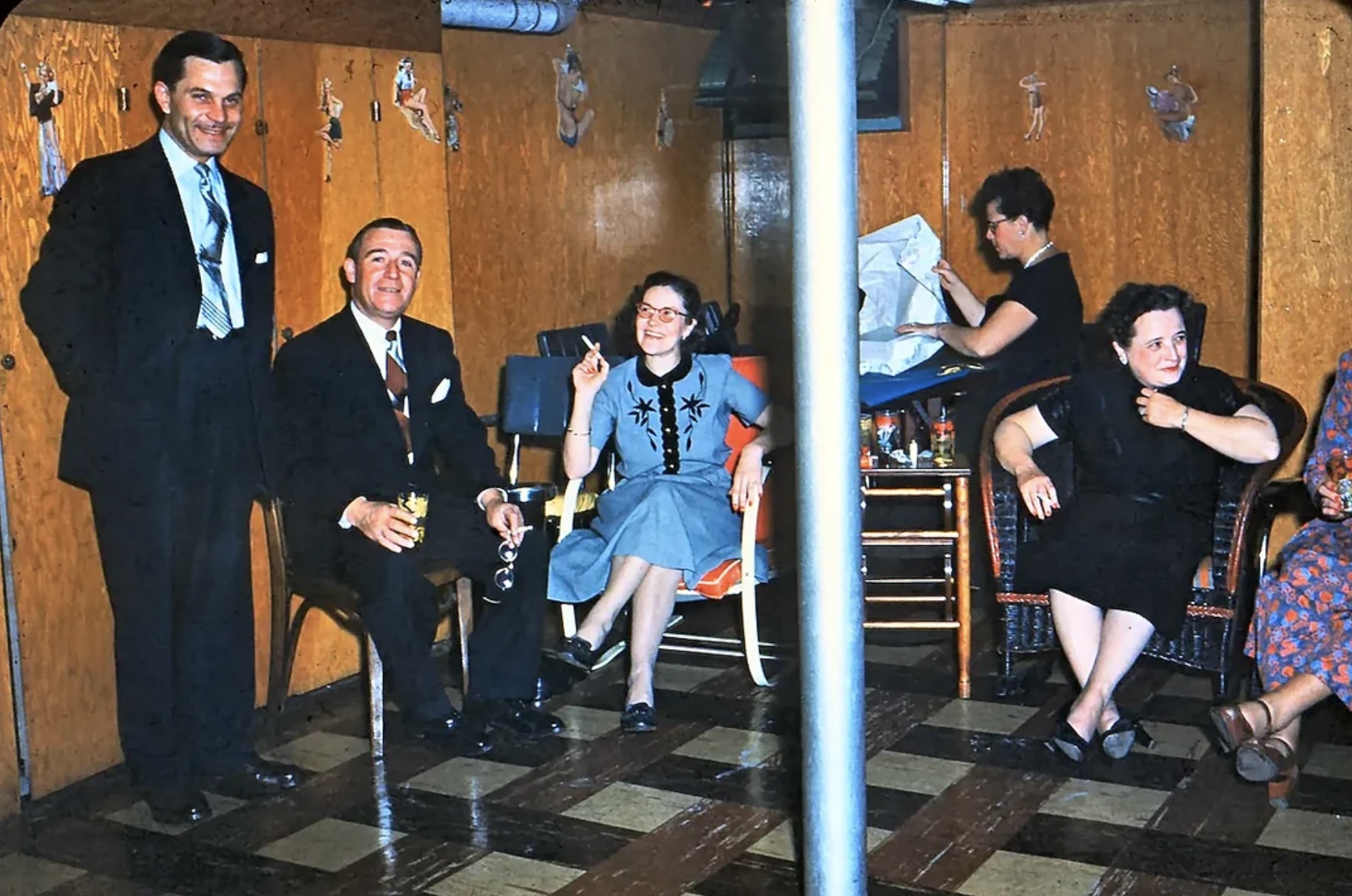 A basement party some time in the early or mid 1950s. Plywood walls, support pole in the center of the room and pinups on the wall. 